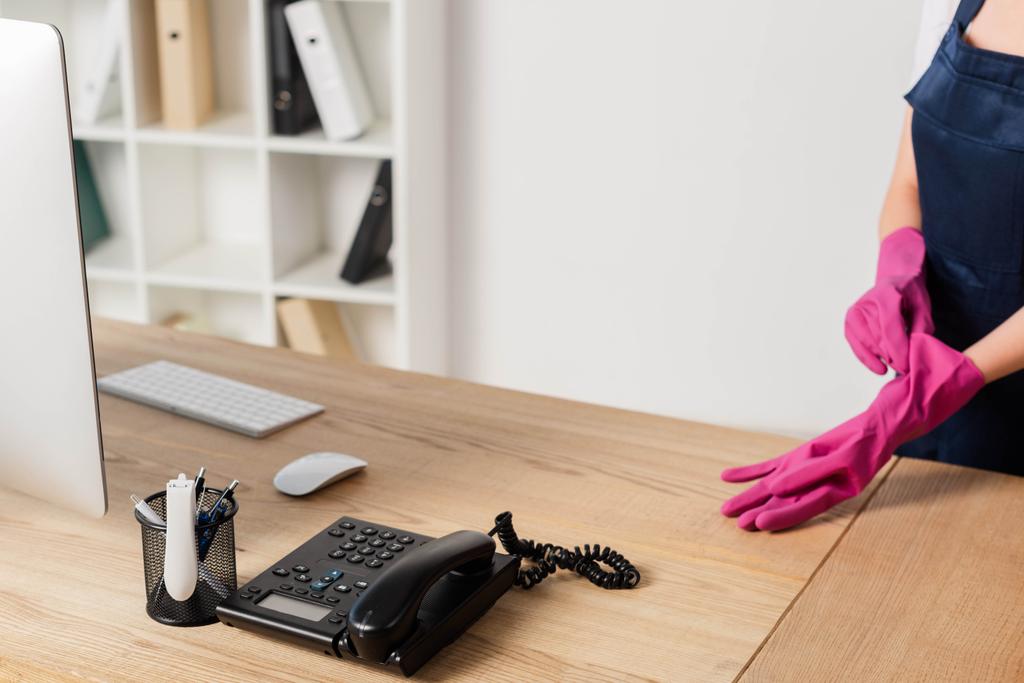 Gekropte weergave van schoner met rubberen handschoen in de buurt van computer en telefoon op kantoor tafel   - Foto, afbeelding