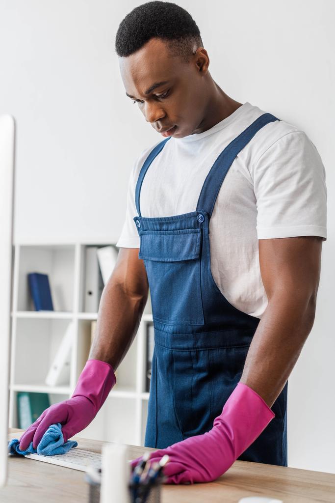 Selective focus of african american cleaner in overalls cleaning computer keyboard in office  - Photo, Image