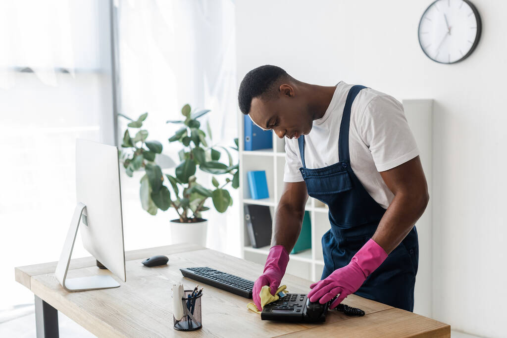 Afro-Amerikaanse werknemer van het schoonmaken van de dienst met behulp van doek, terwijl het schoonmaken van telefoon in de buurt van computer op kantoor tafel  - Foto, afbeelding
