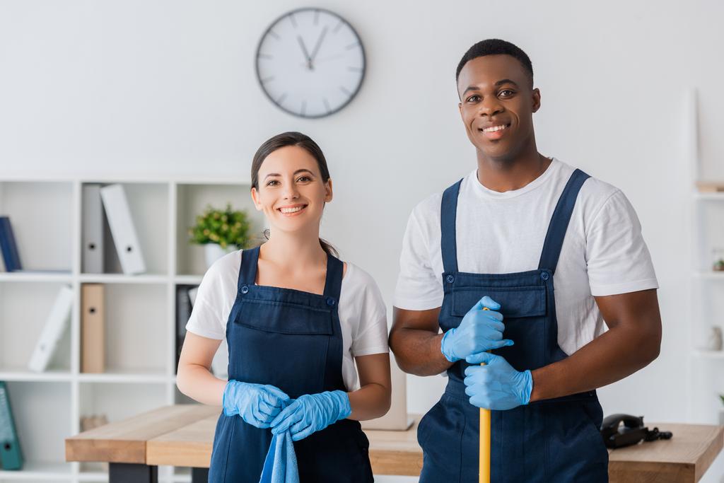 Trabajadores multiétnicos sonrientes del servicio de limpieza sosteniendo trapo y fregona mientras trabajan en la oficina
  - Foto, imagen