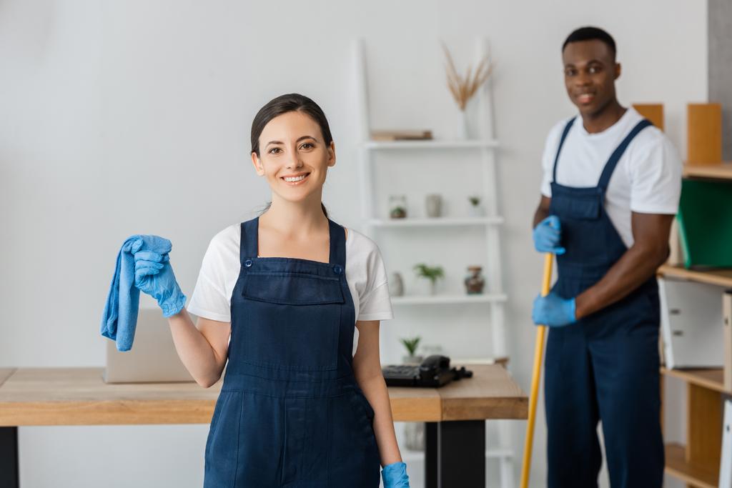 Enfoque selectivo de limpiador sonriente en uniforme sosteniendo trapo cerca de colega afroamericano lavando piso en la oficina
  - Foto, Imagen