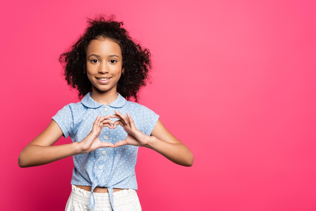 usmívající se roztomilé kudrnaté africké americké dítě ukazující srdce s rukama izolované na růžové - Fotografie, Obrázek