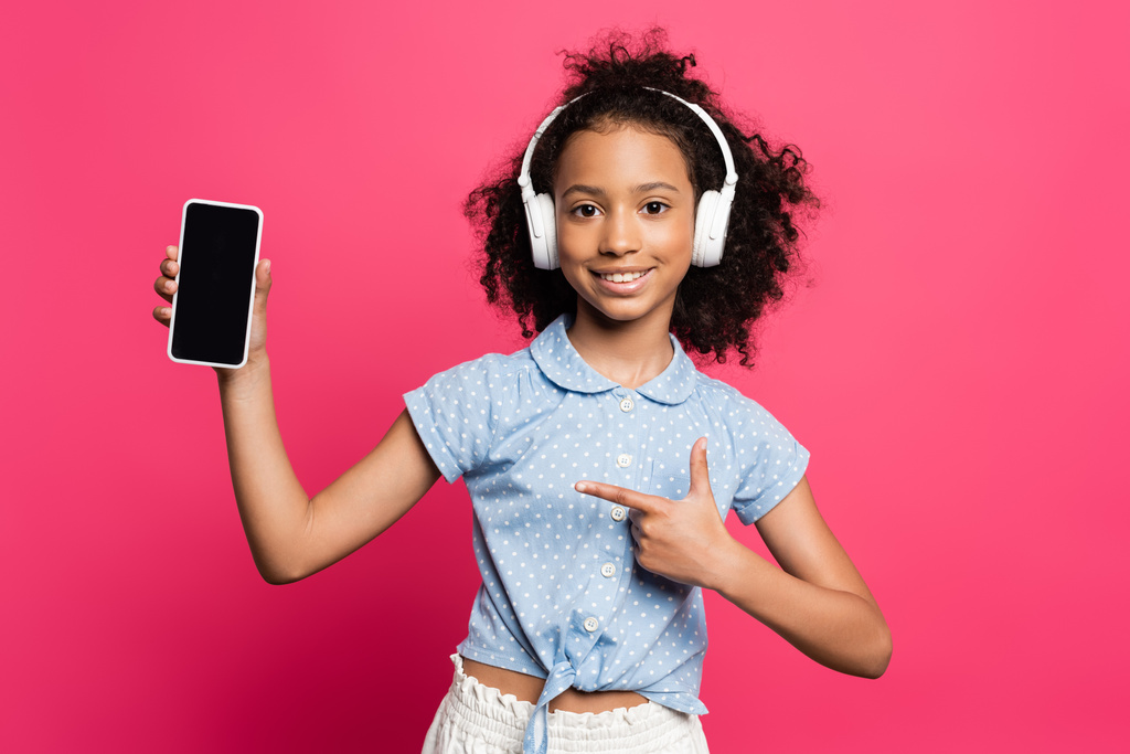 smiling curly african american kid in headphones pointing at smartphone isolated on pink - Photo, Image