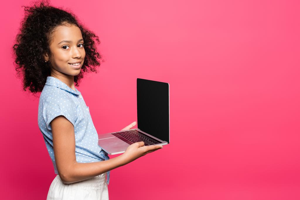 sorridente carino riccio bambino africano americano che tiene il computer portatile con schermo bianco isolato su rosa - Foto, immagini