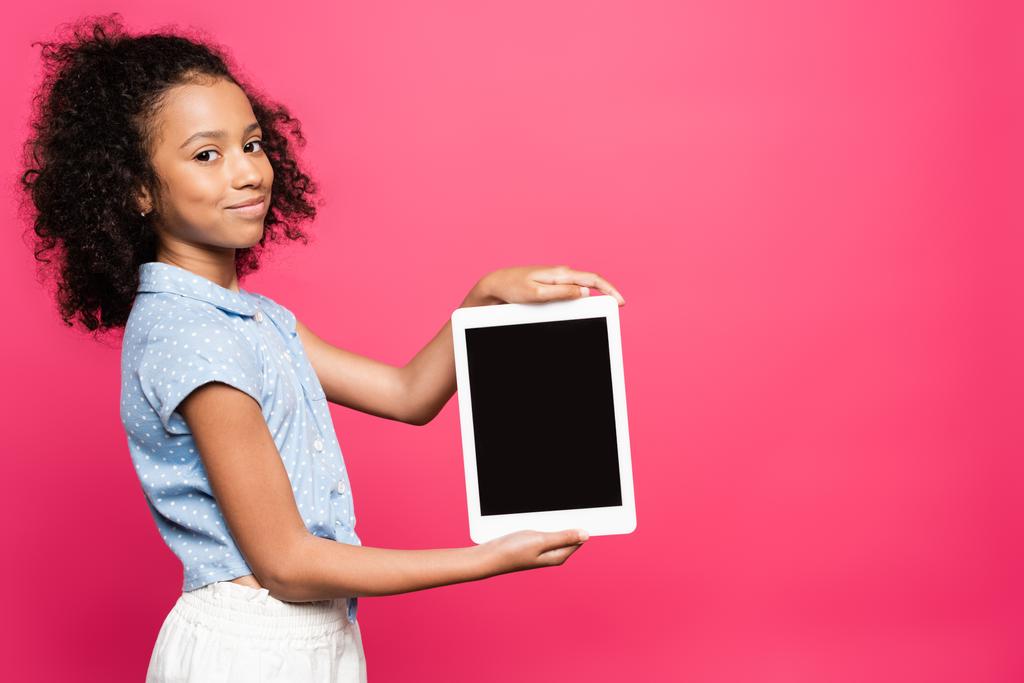 Gülümseyen kıvırcık saçlı Afrikalı Amerikalı çocuk pembe üzerinde izole edilmiş dijital tablet sunuyor. - Fotoğraf, Görsel