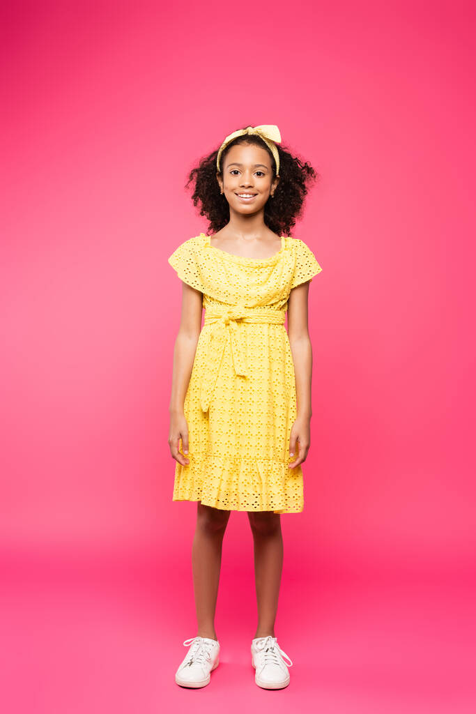 full length view of smiling curly african american child in yellow outfit on pink background - Photo, Image