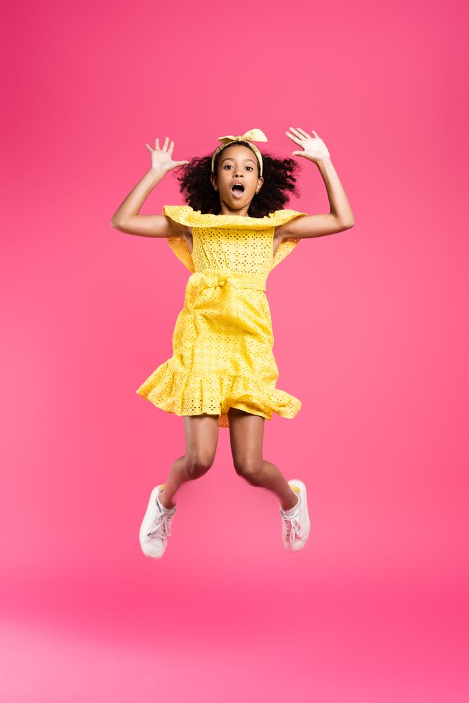 vista completa de niño afroamericano rizado impactado en traje amarillo saltando sobre fondo rosa
 - Foto, imagen