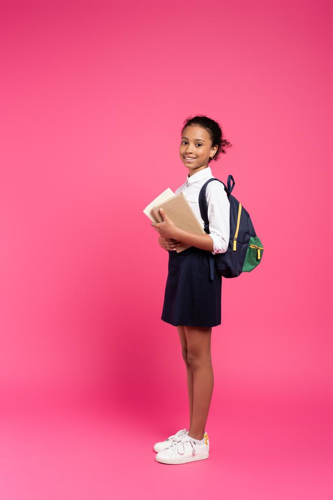 vista lateral de la sonriente colegiala afroamericana con mochila y libros sobre fondo rosa
 - Foto, Imagen