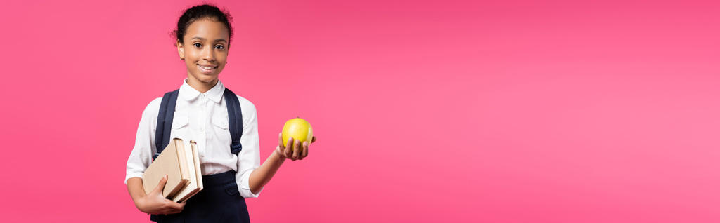 mosolygós afro-amerikai iskolás lány hátizsák és alma elszigetelt rózsaszín, panoráma lövés - Fotó, kép