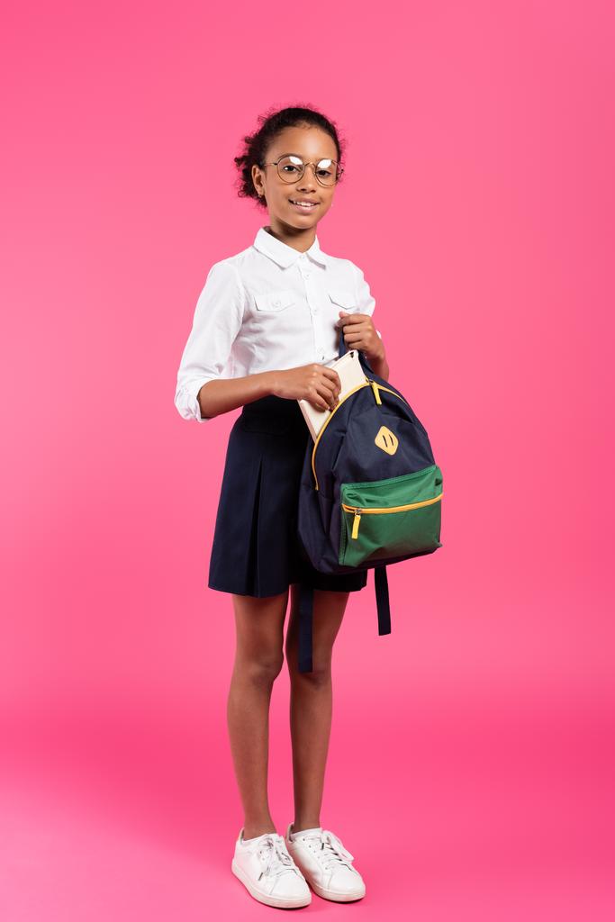 smiling african american schoolgirl in glasses putting book in backpack on pink - Photo, Image