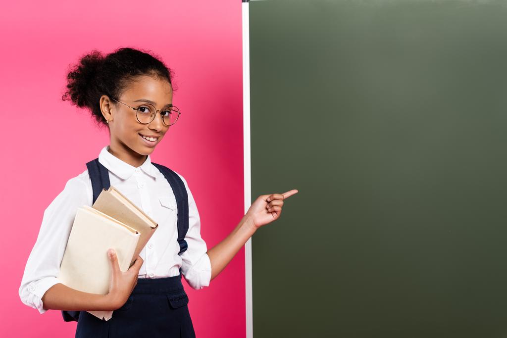 usmívající se africká americká školačka s knihami ukazujícími na prázdné tabuli na růžovém pozadí - Fotografie, Obrázek