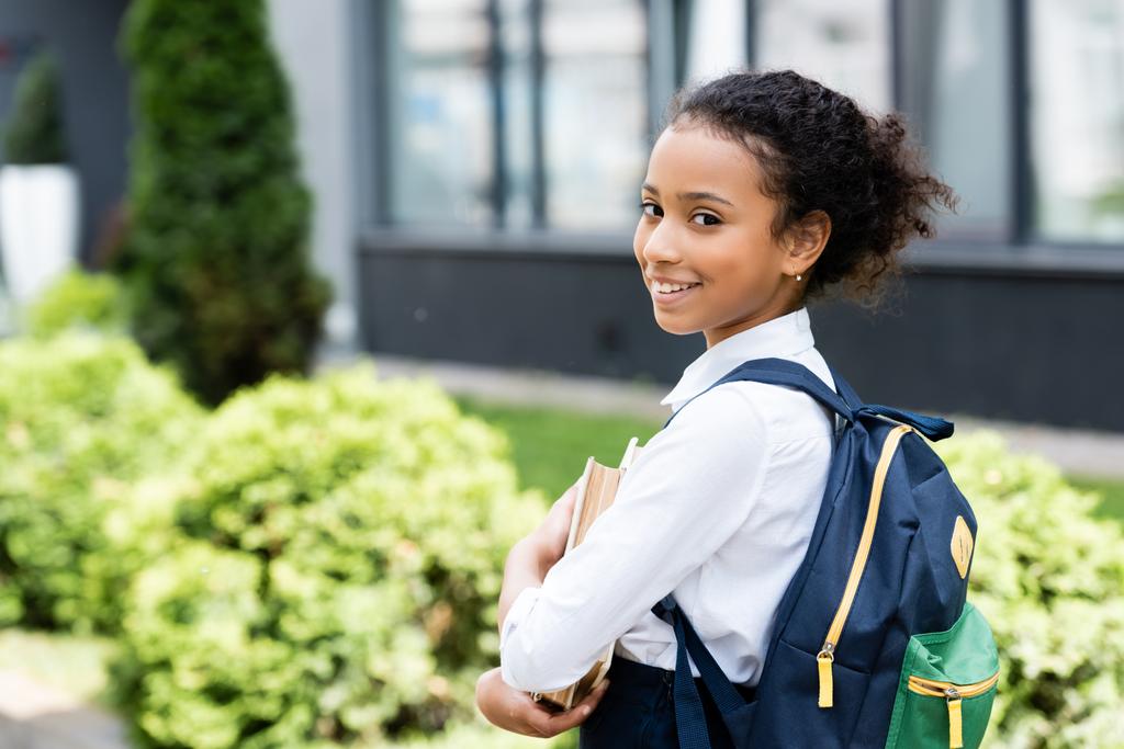 side view of smiling african american schoolgirl with book outdoors - Photo, Image
