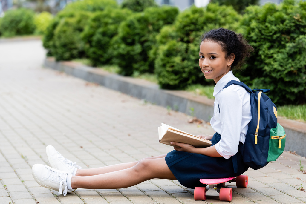 side view of smiling african american schoolgirl reading book while sitting on penny board - Photo, Image