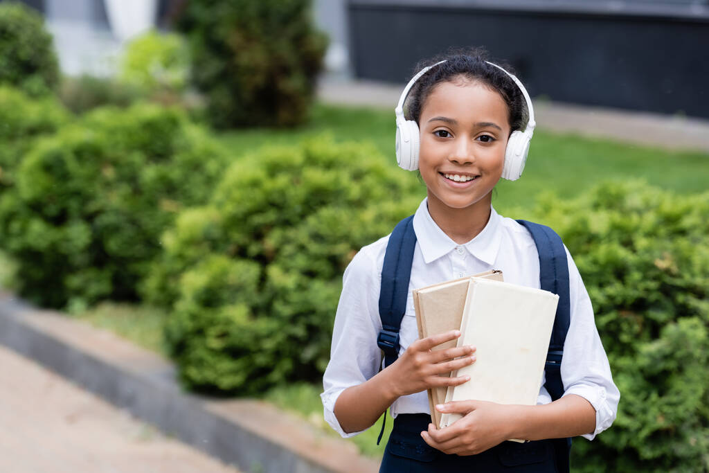 smiling african american schoolgirl in headphones with backpack and books outdoors - Photo, Image