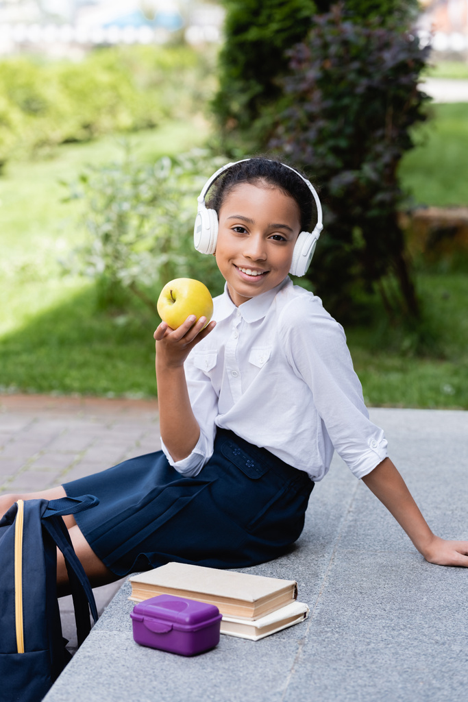 sorrindo Africano americano estudante em fones de ouvido segurando maçã perto de mochila, livros e lancheira ao ar livre
 - Foto, Imagem