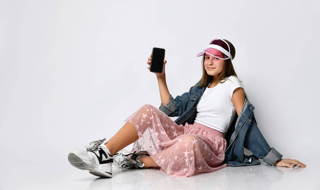 Χαρούμενη γυναίκα που κάθεται στο πάτωμα και δείχνει μια κενή οθόνη smartphone σε ένα ελαφρύ φόντο - Φωτογραφία, εικόνα