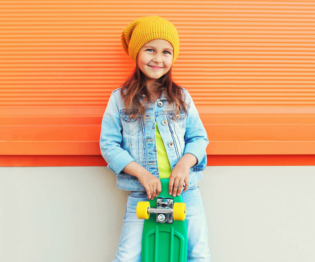 Portrét stylové usmívající se holčička dítě s skateboard na ulici přes oranžové zdi pozadí - Fotografie, Obrázek