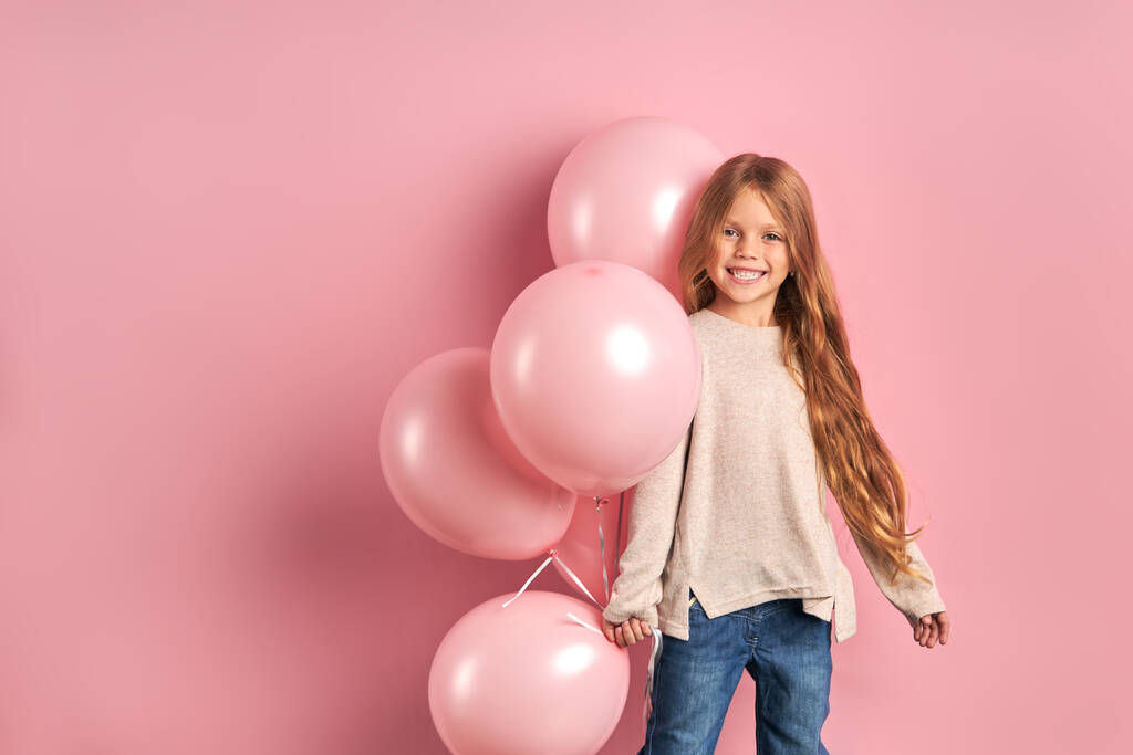 Retrato de niña positiva sobre fondo rosa, sosteniendo racimo de globos de aire rosa
 - Foto, Imagen