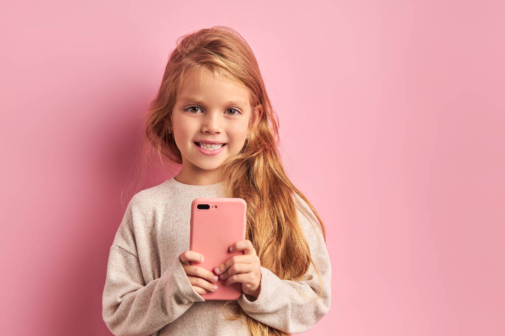 Usmívající se sladká holčička s dlouhými vlasy používat smartphone na růžovém pozadí - Fotografie, Obrázek