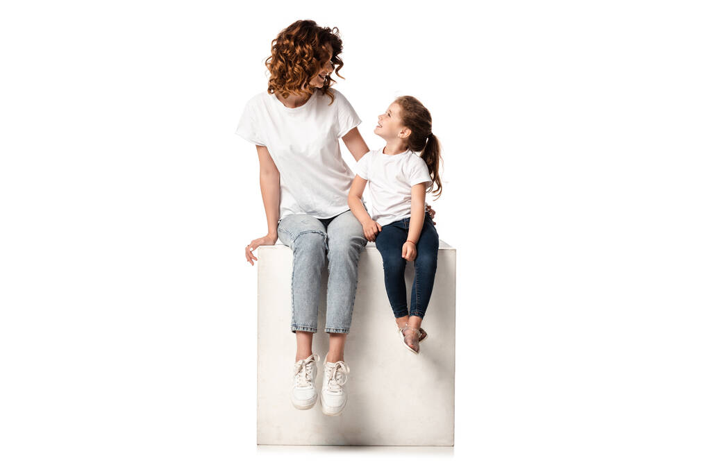 happy mother and daughter looking at each other while sitting on cube on white - Photo, Image