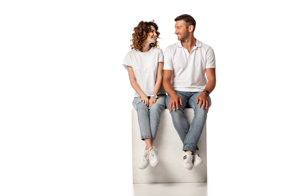 veselý pár usmívá a dívá se na sebe, zatímco sedí na kostce na bílém - Fotografie, Obrázek