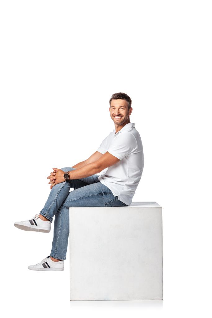 happy man in denim jeans sitting on cube and looking at camera on white - Photo, Image