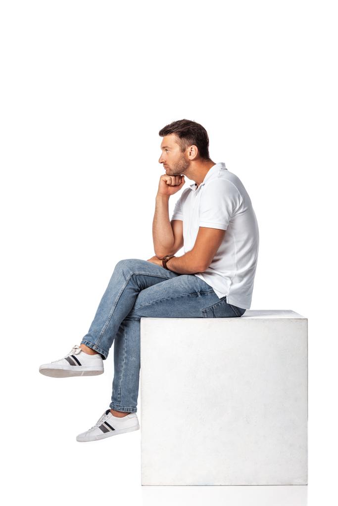 Seitenansicht eines nachdenklichen Mannes in Jeans, der auf einem Würfel auf weißem Grund sitzt - Foto, Bild