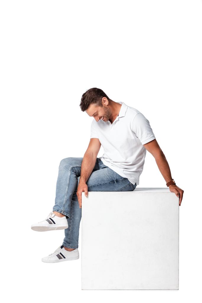 felice uomo in jeans seduto sul cubo e guardando giù isolato su bianco - Foto, immagini