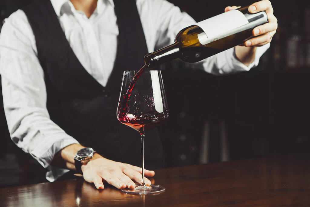 Κοντινό πλάνο του σομελιέ ρίχνει κόκκινο κρασί από το μπουκάλι στο ποτήρι - Φωτογραφία, εικόνα