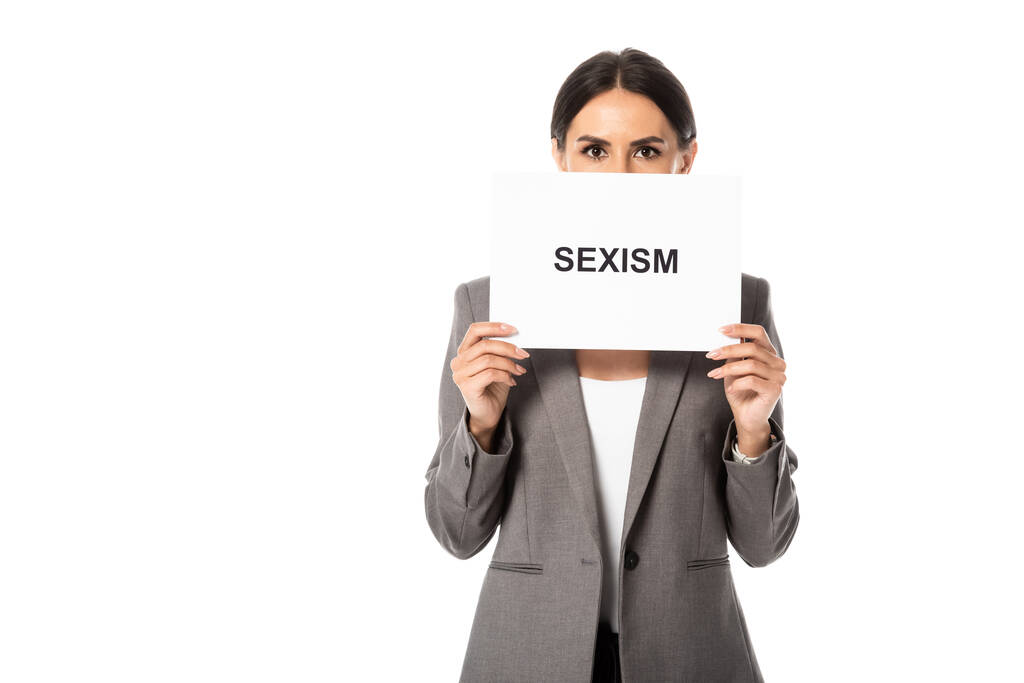 бізнес-леді, що закриває обличчя, тримаючи плакат з написом сексизму і дивлячись на камеру ізольовано на білому
 - Фото, зображення