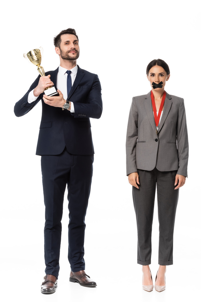 homme d'affaires tenant trophée près d'une femme d'affaires avec ruban adhésif sur la bouche et médaille isolée sur blanc, concept d'inégalité des sexes - Photo, image