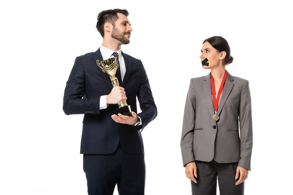 hombre de negocios sosteniendo trofeo de oro y mirando a la mujer de negocios con cinta adhesiva en la boca aislada en blanco
 - Foto, Imagen