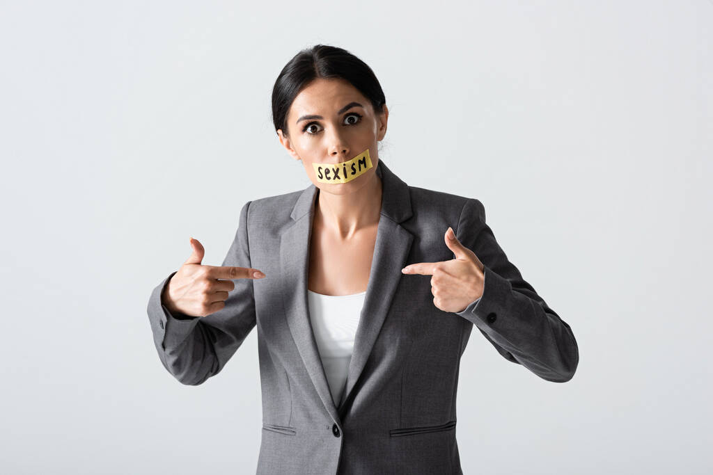mujer de negocios con letras de sexismo en cinta adhesiva mirando a la cámara mientras señala con los dedos a sí misma aislada en blanco
 - Foto, Imagen