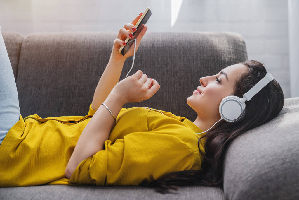Vista lateral de la mujer joven escuchando música en línea en el teléfono inteligente en los auriculares
 - Foto, imagen