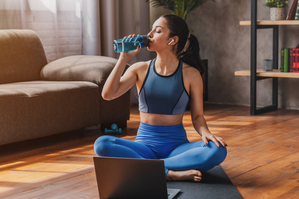 Fit Frau in Sportbekleidung sitzt auf Matte und trinkt aus Wasserflasche, während sie Laptop im heimischen Wohnzimmer benutzt - Foto, Bild