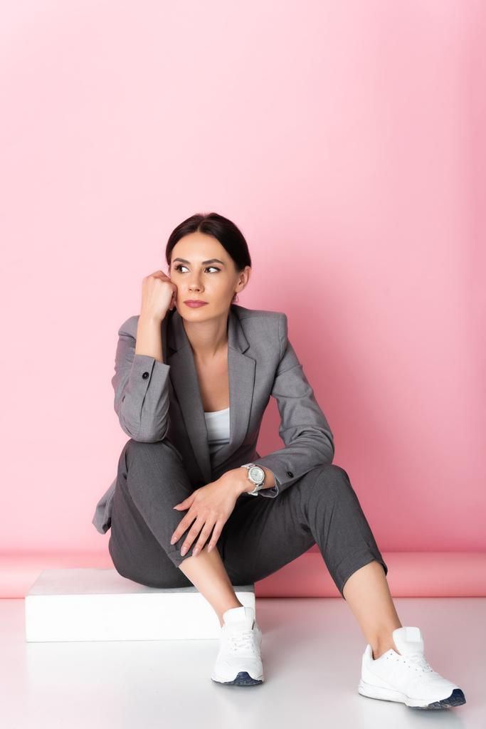 pensive businesswoman in suit and white sneakers looking away while sitting on pink  - Photo, Image