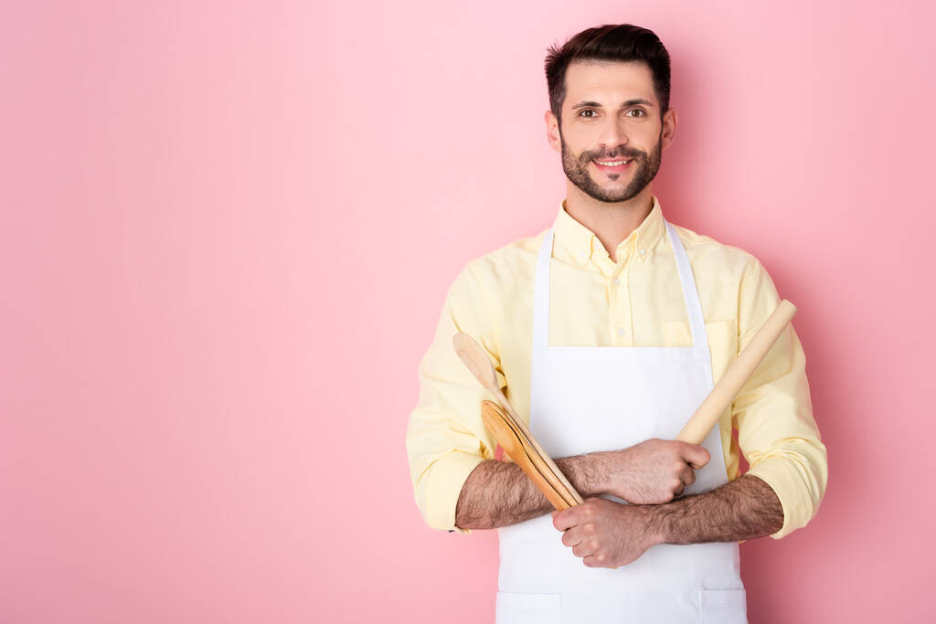 happy bearded man in apron holding wooden rolling pin and spoon on pink  - Photo, Image