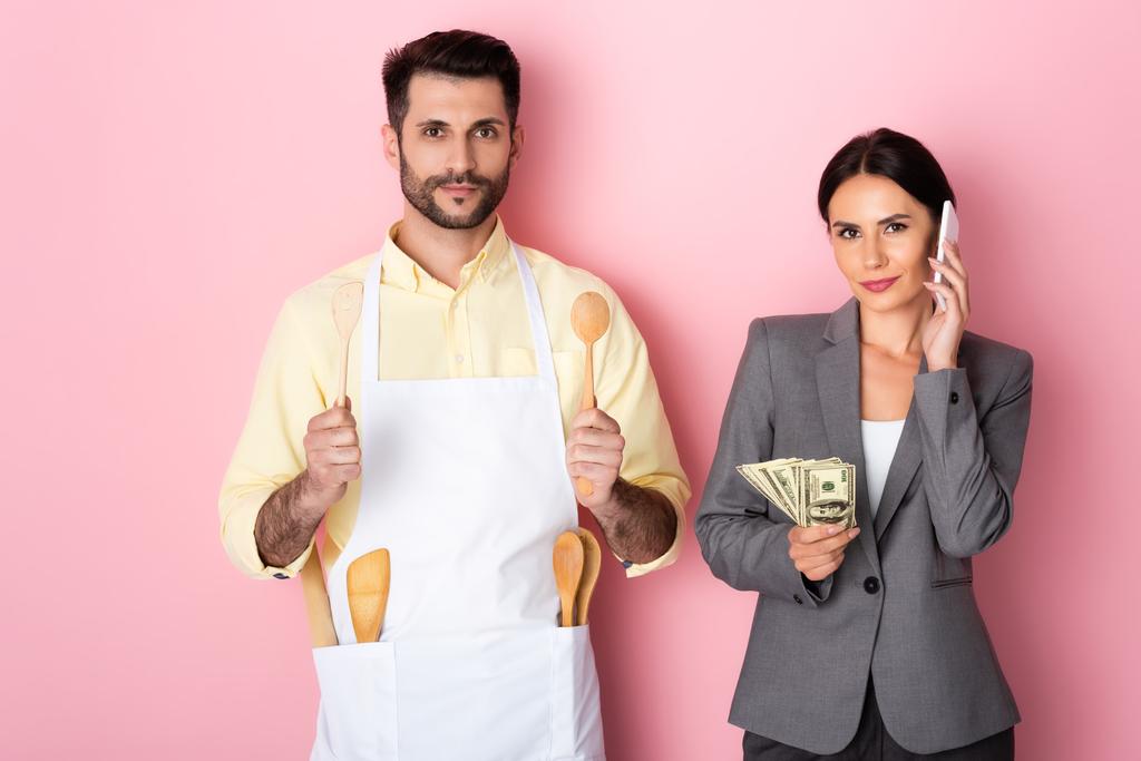 schöner Mann in Schürze mit Kochlöffel und Spachtel in der Nähe Geschäftsfrau mit Geld reden auf Smartphone auf rosa  - Foto, Bild