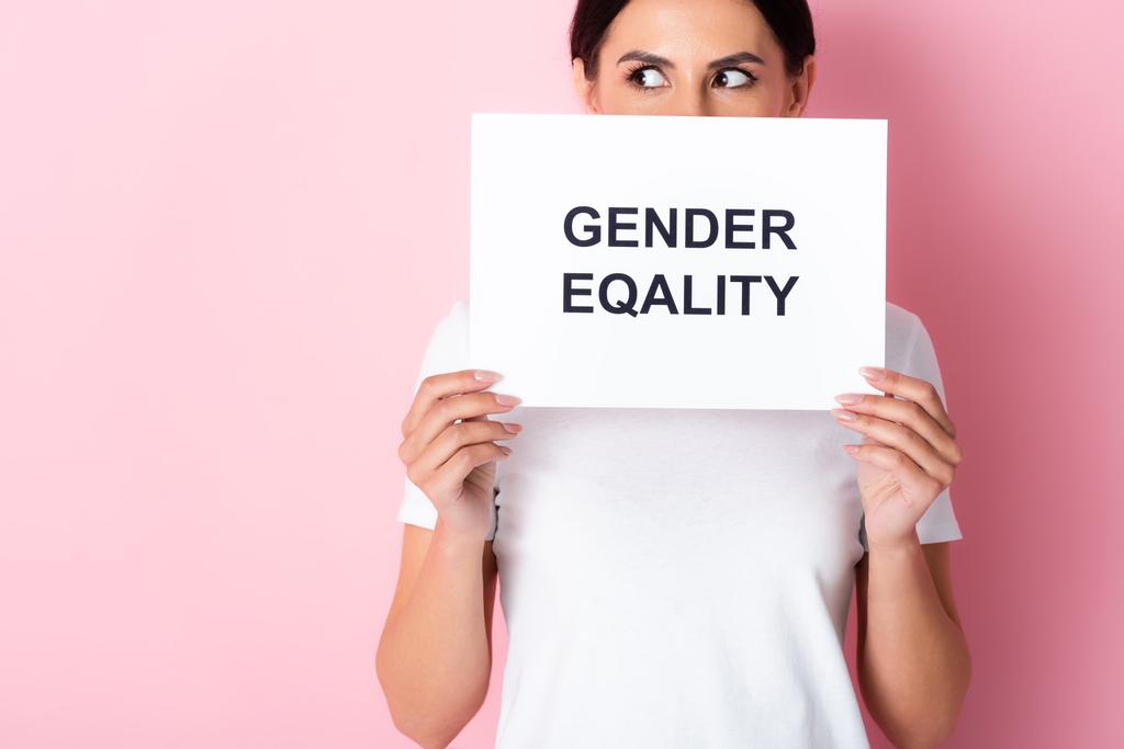женщина в белой футболке покрывает лицо с гендерным равенством надпись на плакате и глядя на розовый
  - Фото, изображение