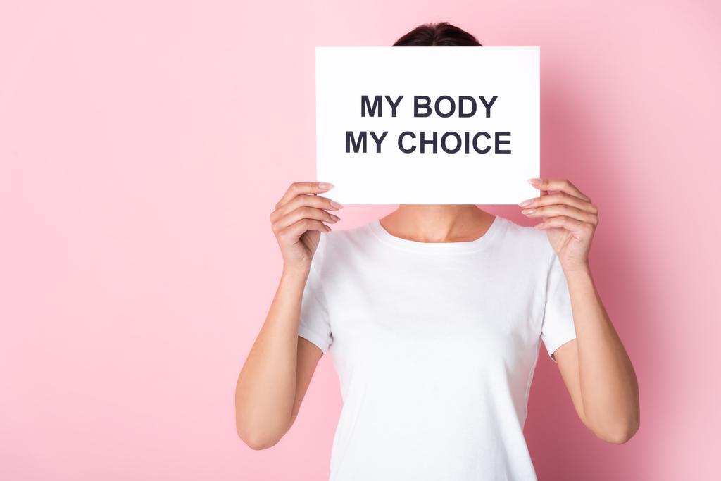 женщина в белой футболке держит плакат с моим телом мой выбор буквы, покрывая лицо на розовый
  - Фото, изображение
