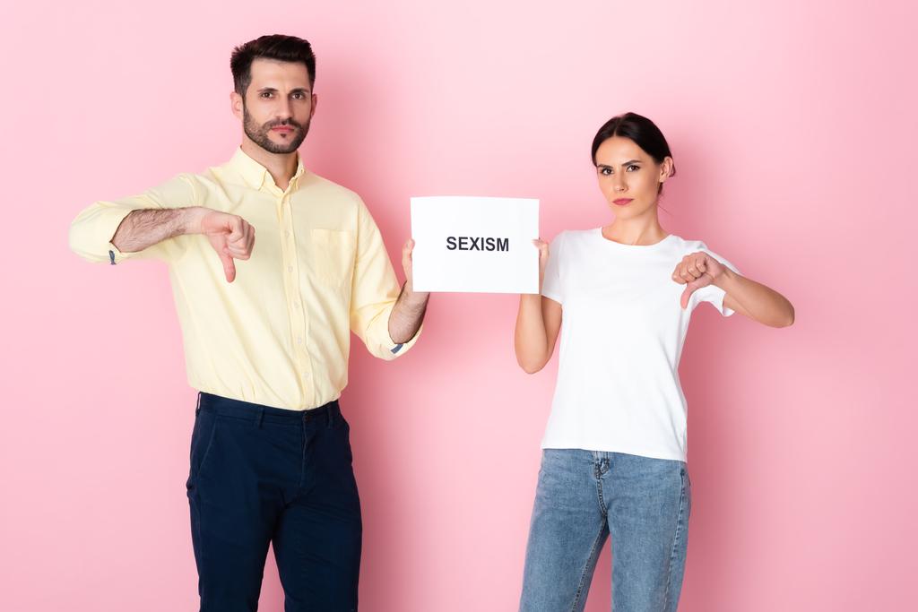 homem e mulher em t-shirt branca segurando cartaz com sexismo lettering e mostrando polegares para baixo em rosa
  - Foto, Imagem