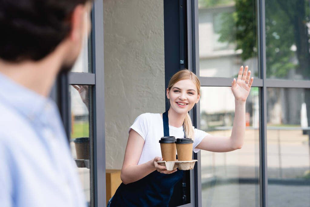 Enfoque selectivo de camarera sonriente sosteniendo vasos de papel y saludando al hombre cerca de la cafetería
  - Foto, imagen