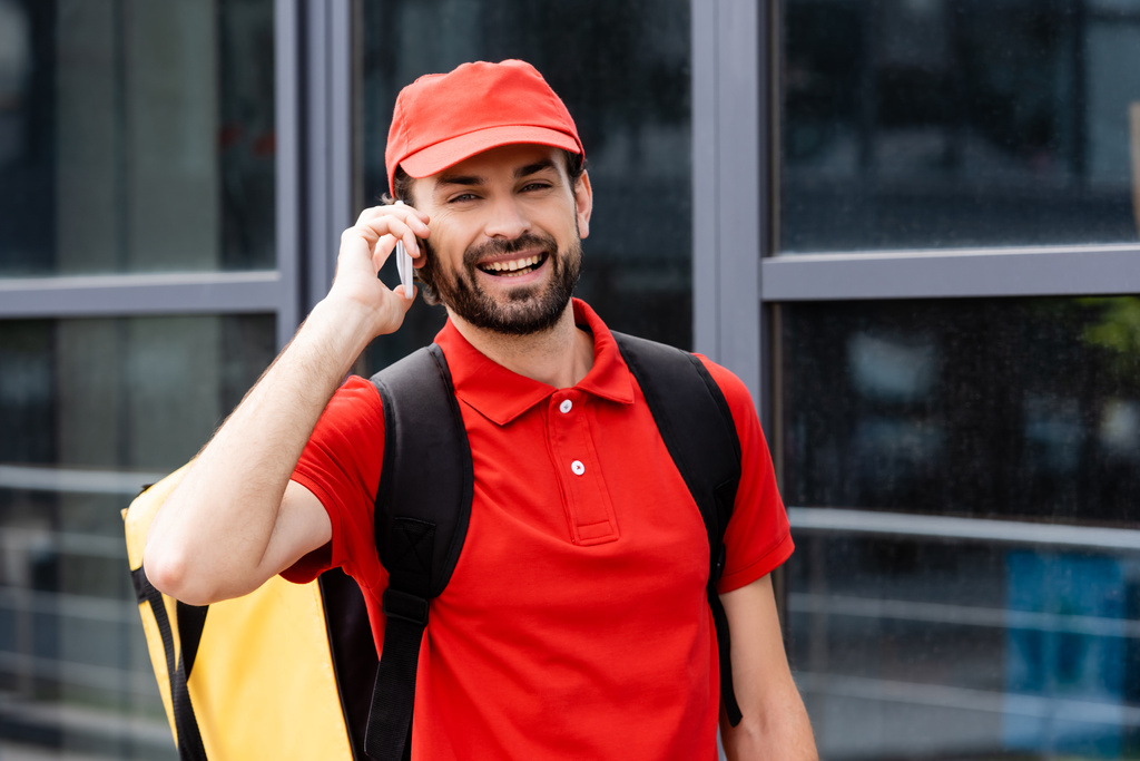 Mensajero sonriente con mochila térmica hablando en smartphone en la calle urbana
  - Foto, Imagen