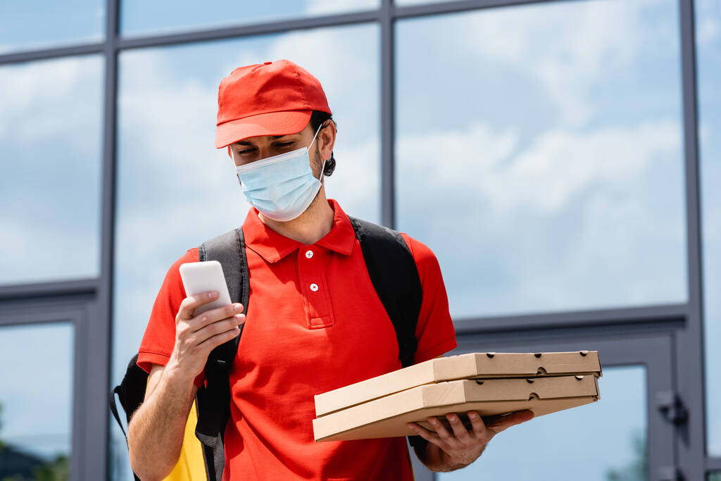 Ταχυδρόμος με ιατρική μάσκα χρησιμοποιώντας smartphone και κρατώντας κουτιά πίτσας σε αστικό δρόμο  - Φωτογραφία, εικόνα