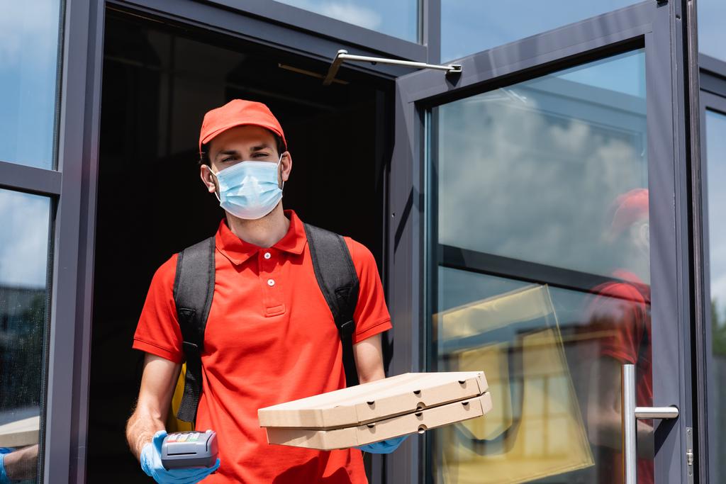 Enfoque selectivo de mensajero en máscara médica que sostiene terminal de pago y cajas de pizza cerca del edificio en la calle urbana
  - Foto, imagen
