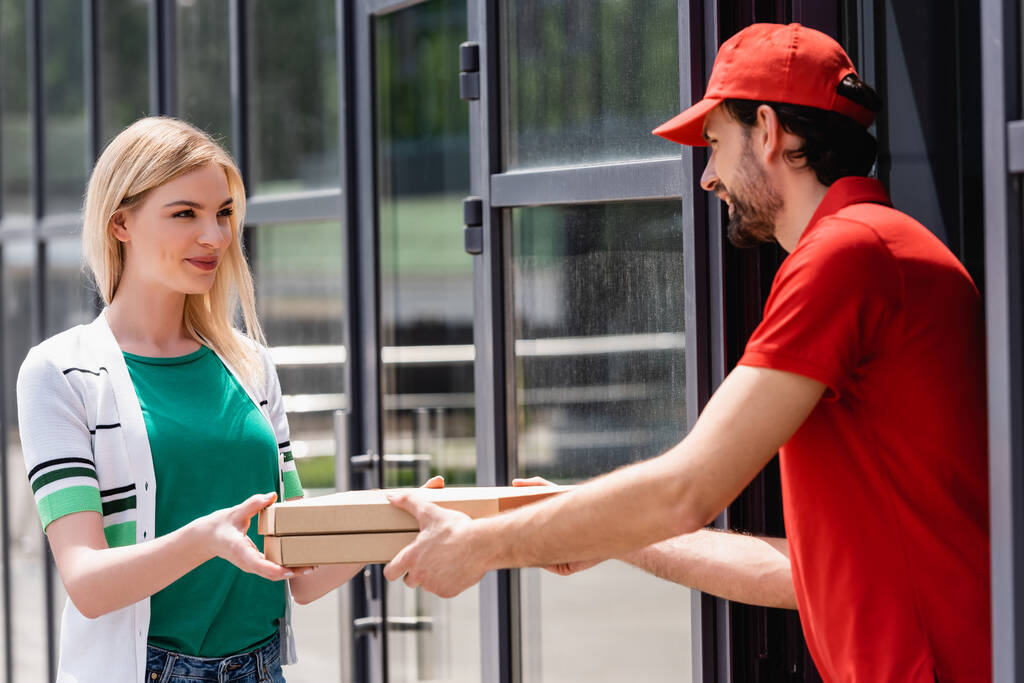 Garçom sorridente em uniforme dando caixas de pizza para jovem mulher perto de café na rua urbana
  - Foto, Imagem