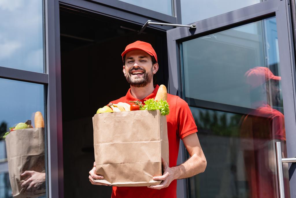 Позитивный курьер держит бумажный пакет со свежими овощами и багетом на городской улице  - Фото, изображение