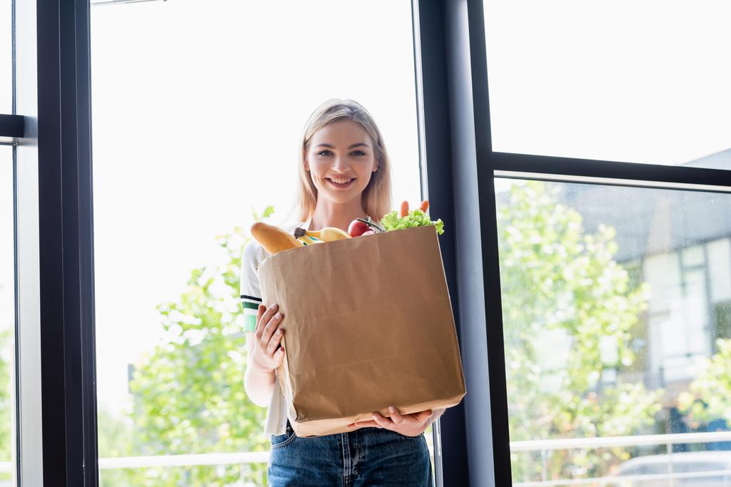 Uśmiechnięta kobieta trzyma torbę na zakupy ze świeżymi warzywami i patrzy w kamerę w domu  - Zdjęcie, obraz