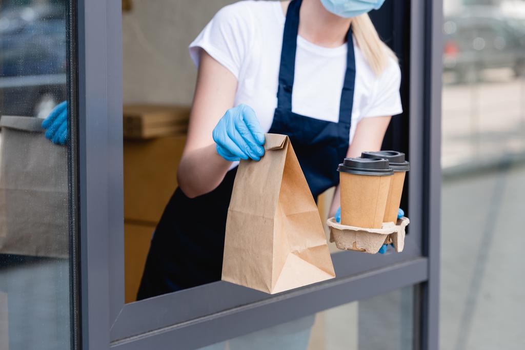 Vista recortada de la camarera en guantes de látex sosteniendo vasos de papel y paquete cerca de la ventana de la cafetería
 - Foto, imagen