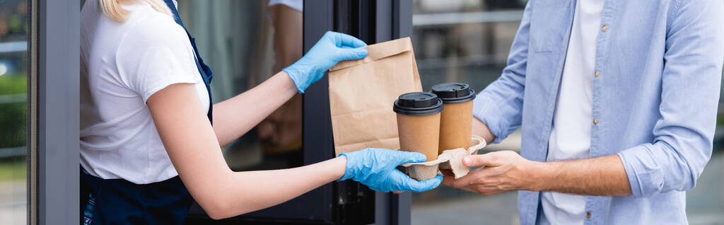 Panoramiczne ujęcie mężczyzny otrzymującego paczkę i papierowe kubki od kelnerki w lateksowych rękawiczkach w pobliżu kawiarni  - Zdjęcie, obraz